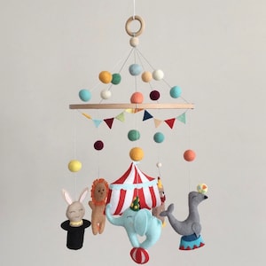 May include: A colorful felt baby mobile with a circus theme. It features a red and white striped tent, a blue elephant, a gray seal, a brown lion, a white rabbit, and colorful felt balls.
