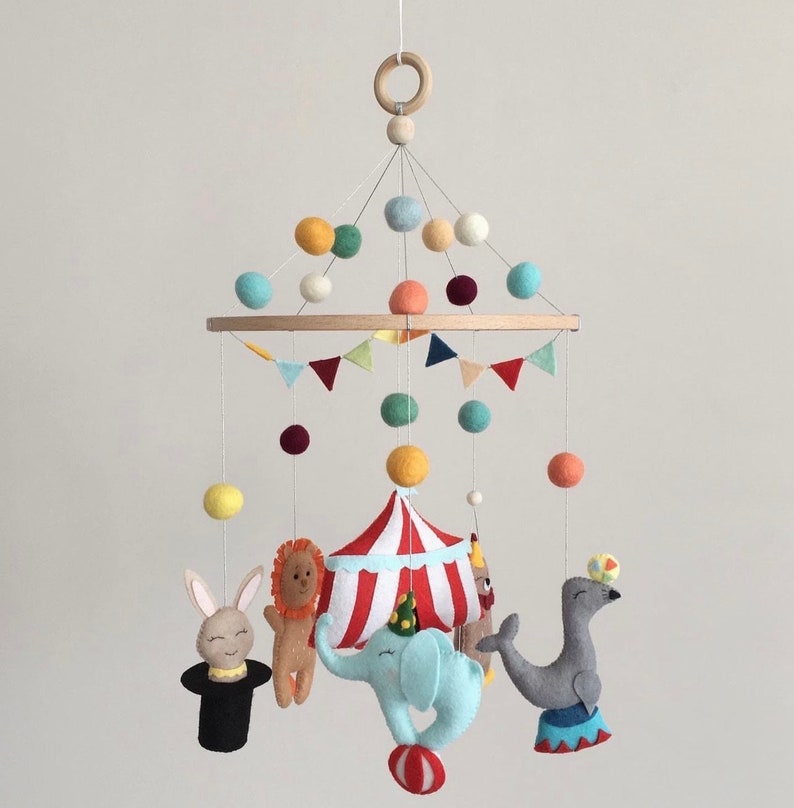 May include: A colorful felt baby mobile with a circus theme. It features a red and white striped tent, a blue elephant, a gray seal, a brown lion, a white rabbit, and colorful felt balls.