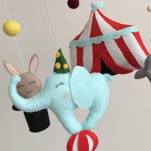 May include: A baby mobile with a blue elephant balancing a red and white ball, a brown rabbit in a black top hat, a red and white striped tent, and yellow and maroon felt balls.