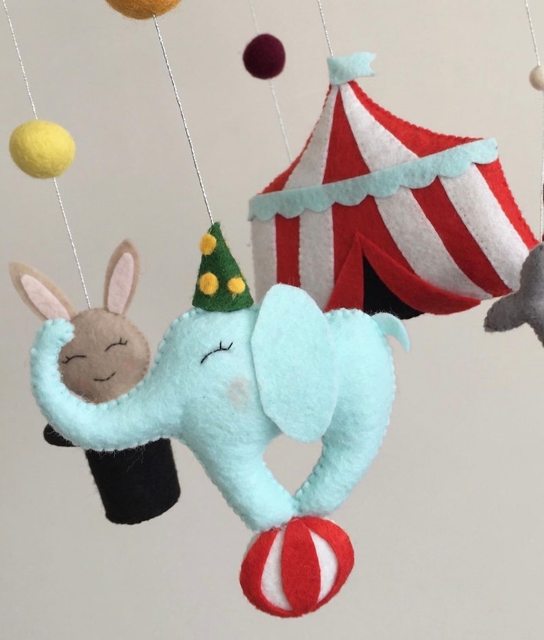 May include: A baby mobile with a blue elephant balancing a red and white ball, a brown rabbit in a black top hat, a red and white striped tent, and yellow and maroon felt balls.