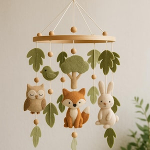 May include: A baby mobile with a woodland theme, featuring felt animals and foliage. The mobile includes a light brown owl, a fox, a rabbit, a bird, a tree, and green leaves, suspended from a wooden ring with wooden beads. The design is soft and neutral.