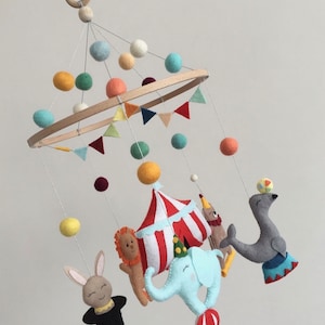 May include: A colorful felt baby mobile with a circus theme. It features a red and white striped tent, a blue elephant, a gray seal balancing a ball, a lion, a rabbit with a top hat, and colorful felt balls.
