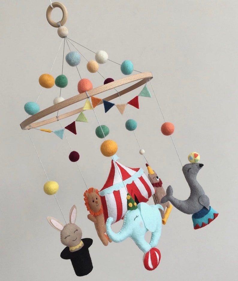 May include: A colorful felt baby mobile with a circus theme. It features a red and white striped tent, a blue elephant, a gray seal balancing a ball, a lion, a rabbit with a top hat, and colorful felt balls.