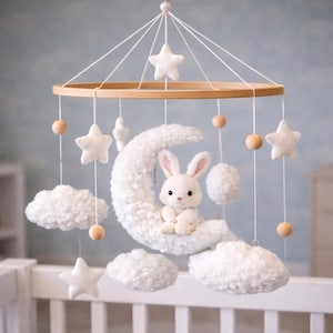 Puede incluir: Móvil para bebé con un conejito blanco de peluche sobre una luna creciente, rodeado de estrellas blancas, nubes y cuentas de madera. Colgando de un aro de madera, es una encantadora decoración para la habitación del bebé.