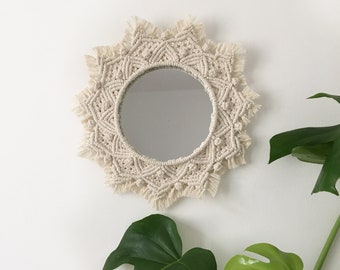 Peut inclure: Un miroir rond avec un cadre en macramé blanc. Le cadre est fait de corde nouée et a une frange autour du bord. Le miroir est suspendu à un mur blanc.