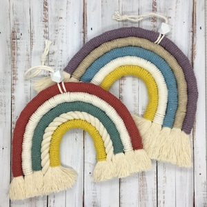 Könnte beinhalten: Zwei bunte Makramee-Regenbögen mit weißen Quasten, die an einer weißen Holzwand hängen. Der Regenbogen links hat rote, weiße, grüne, gelbe und weiße Streifen. Der Regenbogen rechts hat lila, beige, blaue, gelbe und weiße Streifen.