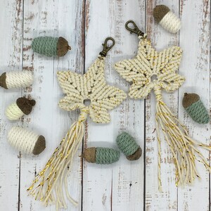 Op de afbeelding: Twee witte macramé ster-sleutelhangers met gouden accenten en kwastjes die hangen aan een witte houten achtergrond met kleine groene en bruine eikeldecoraties.