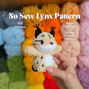 Op de afbeelding: Een gehaakt lynx-speeltje in beige, zwart en wit, gehouden voor een kleurrijke garenachtergrond. De tekst "No Sew Lynx Pattern" en extra details worden weergegeven. De lynx heeft zwarte oren en vlekken.