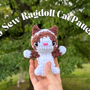 May include: A handmade, no-sew Ragdoll cat pattern. The plush cat is brown, white, and pink, with large black eyes. The cat is held in a hand against a blurred green background. The text "No-Sew Ragdoll Cat Pattern" is arched above the cat.