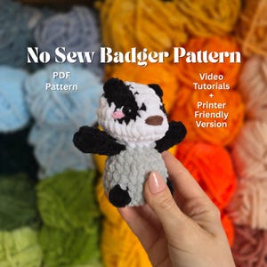 Puede incluir: Un peluche de tejón tejido a ganchillo con detalles en negro, blanco y gris. El peluche tiene una nariz rosa y un hocico marrón. La imagen incluye el texto "No Sew Badger Pattern" y "PDF Pattern". Texto adicional dice "Video Tutorials + Printer Friendly Version".