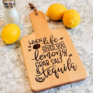 May include: A wooden cutting board with a handle and the engraved text "When life gives you lemons grab salt & tequila." The board is accompanied by three yellow lemons and a glass salt shaker.