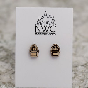 May include: Wooden cactus stud earrings. Each earring features a black outline of a cactus in a pot. The earrings are displayed on a white card with the text "NWC NORTH WEST CREATIVE" and a mountain logo.