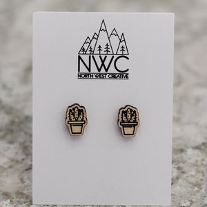 May include: A pair of wooden cactus earrings on a white card. The earrings are light brown with a dark brown outline and feature a cactus in a pot design. The card has the text "NWC NORTH WEST CREATIVE" with a mountain and tree logo.