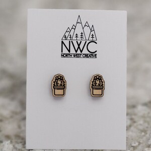 May include: Two wooden cactus stud earrings on a white card. The earrings are light brown with a black outline of a cactus in a pot. The card has a mountain and tree logo with the text "NWC NORTH WEST CREATIVE".