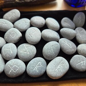 May include: A collection of twenty-four smooth, grey river stones arranged on a black velvet pouch. Each stone features a unique, hand-carved white rune symbol etched into its surface. The stones are oval and rounded in shape, displaying natural variations in texture and grey tones across the set.