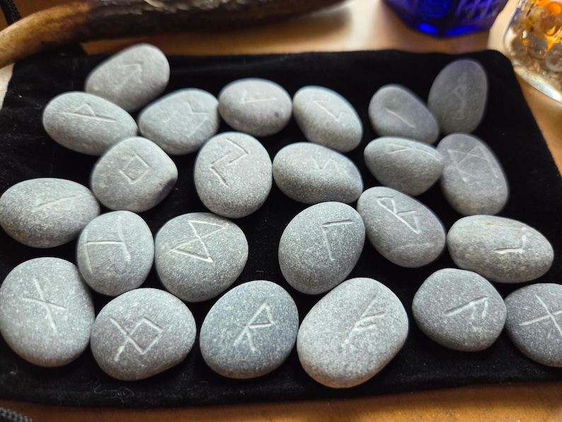 May include: A collection of twenty-four smooth, grey river stones arranged on a black velvet pouch. Each stone features a unique, hand-carved white rune symbol etched into its surface. The stones are oval and rounded in shape, displaying natural variations in texture and grey tones across the set.