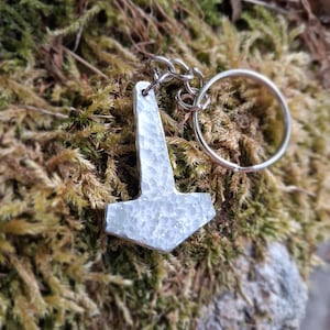May include: A silver-colored metal keychain featuring a hammer-shaped pendant with a textured, hammered surface. The pendant is attached to a circular metal split ring by a small jump ring, creating a functional accessory for keys or bags.