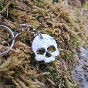 Könnte beinhalten: Ein silberfarbener Metallschlüsselanhänger mit einem totenkopfförmigen Anhänger, der eine gehämmerte, strukturierte Oberfläche aufweist. Der Totenkopfanhänger ist mit einem kurzen silberfarbenen Metallkettenglied an einem runden Metallring befestigt.