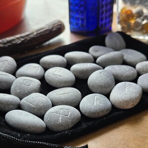 May include: A collection of smooth, grey river stones engraved with various runic symbols, arranged on a black velvet drawstring pouch. Each stone features a single carved character, showcasing natural textures and variations in shape and size.