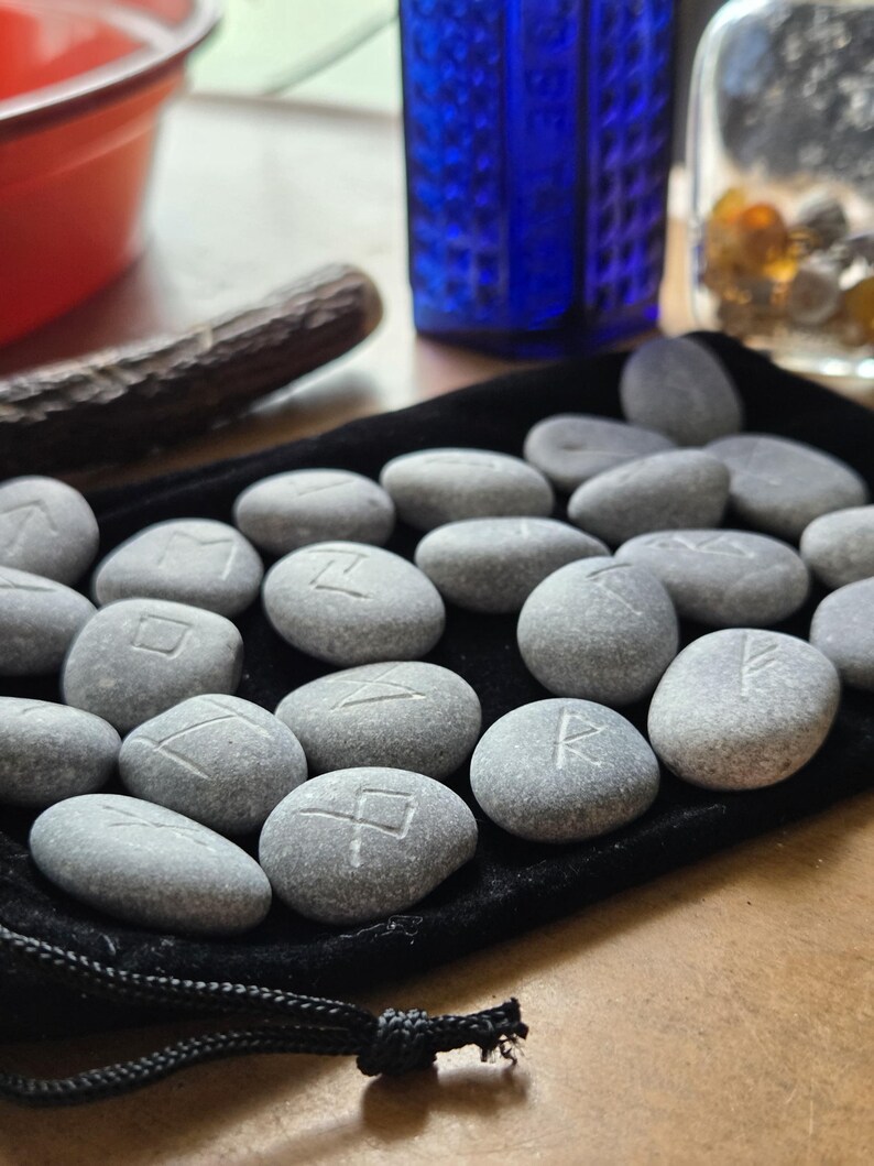 May include: A collection of smooth, grey river stones engraved with various runic symbols, arranged on a black velvet drawstring pouch. Each stone features a single carved character, showcasing natural textures and variations in shape and size.