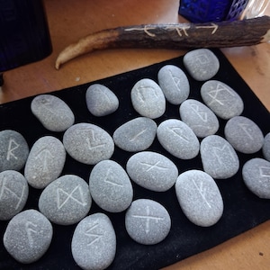 May include: A collection of smooth, grey river stones engraved with white runic symbols, arranged on a rectangular black velvet mat. A single piece of dark brown antler with carved white markings lies horizontally above the stones.