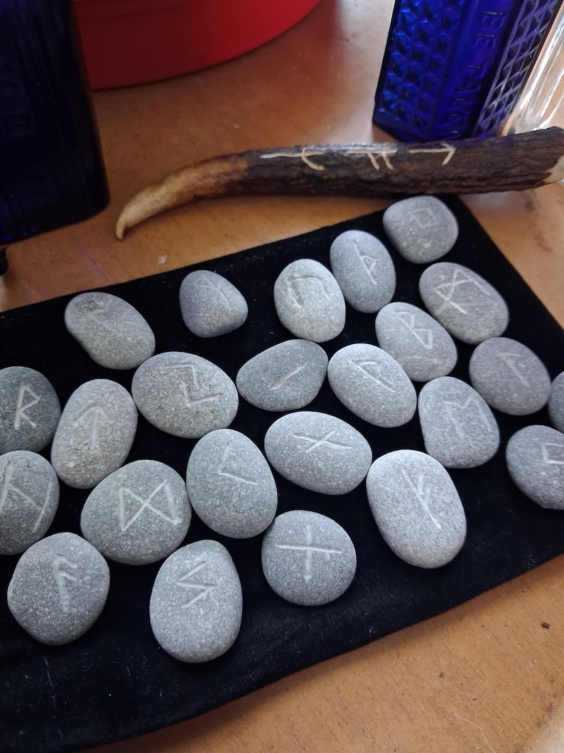 May include: A collection of smooth, grey river stones engraved with white runic symbols, arranged on a rectangular black velvet mat. A single piece of dark brown antler with carved white markings lies horizontally above the stones.
