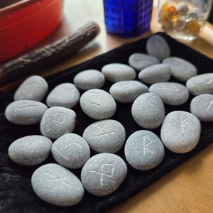 May include: A collection of smooth, grey river stones featuring etched runic symbols, arranged on a black velvet drawstring pouch. Each stone is oval-shaped with a matte texture, displaying distinct carved characters across the surface.