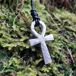 May include: A silver-colored Ankh pendant with a textured surface hangs from a black cord. The pendant is suspended against a backdrop of green moss and foliage, creating a natural aesthetic.