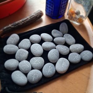 May include: A collection of smooth, grey river stones featuring etched white runic symbols, arranged on a black velvet drawstring pouch. Each stone displays a unique character, with the set spread across the fabric surface in a loose formation.
