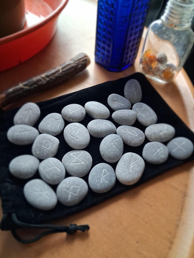 May include: A collection of smooth, grey river stones featuring etched white runic symbols, arranged on a black velvet drawstring pouch. Each stone displays a unique character, with the set spread across the fabric surface in a loose formation.