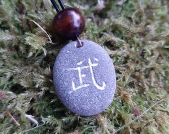 Puede incluir: Colgante de piedra gris ovalada con un carácter japonés blanco. Cuelga de un cordón negro con una cuenta de madera marrón oscuro. El fondo es musgo verde.