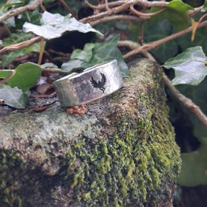 May include: A silver-toned metal ring with a wide, hammered band texture. The ring features a small, dark, engraved phoenix emblem centered on the outer surface. The band has an open design, showing a gap at the top of the ring's circumference.