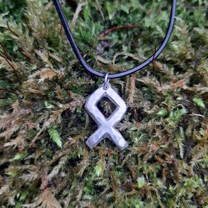 May include: A silver pendant necklace with a Viking rune symbol on a black leather cord. The pendant is on a bed of green moss.