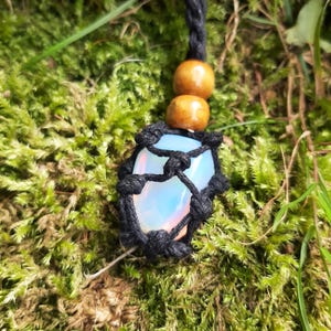 May include: A handcrafted necklace with a light blue crystal pendant in a black woven net. The necklace includes two wooden beads and a black cord. The background is green moss.
