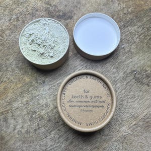 May include: Open round cardboard container with green powder, next to an empty lid and a container with text: "for teeth & gums" and "0.5 ounces". The product is a mineral and organic herbal toothpaste powder.