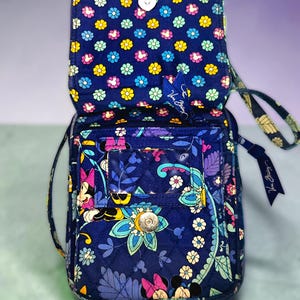 May include: A navy blue quilted crossbody bag featuring a colorful floral and Minnie Mouse pattern. The bag has a fold-over flap with a magnetic snap closure, a clear plastic ID window on the front, and a wrist strap with a Vera Bradley logo tag.