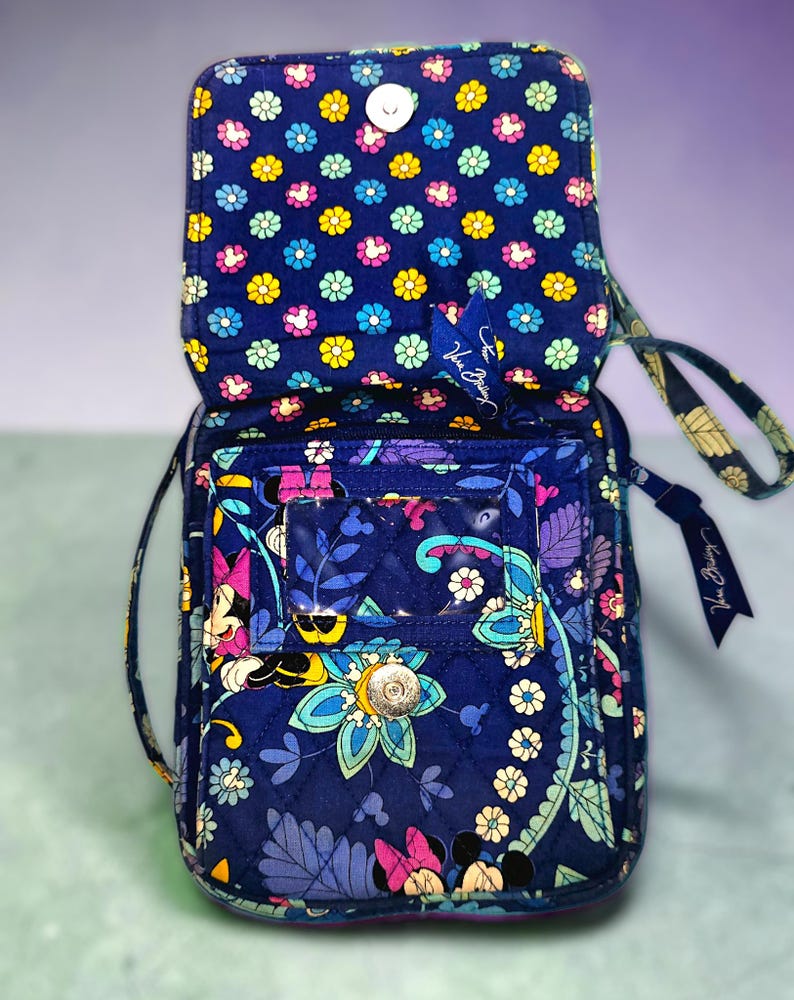 May include: A navy blue quilted crossbody bag featuring a colorful floral and Minnie Mouse pattern. The bag has a fold-over flap with a magnetic snap closure, a clear plastic ID window on the front, and a wrist strap with a Vera Bradley logo tag.