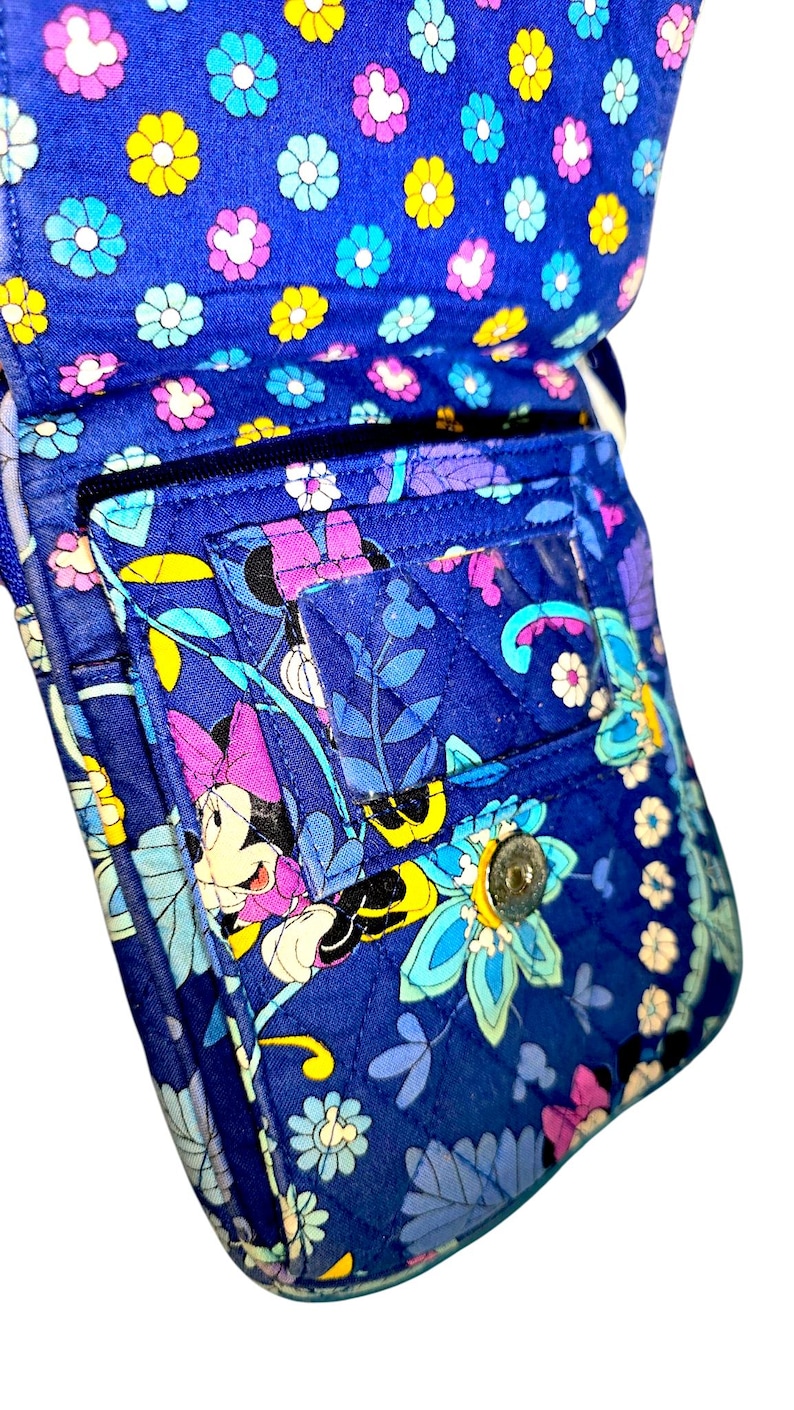 May include: A quilted fabric crossbody bag in a vibrant blue featuring a repeating pattern of colorful flowers and Minnie Mouse characters. The bag includes a front flap with a clear plastic ID window, a metal snap closure, and a zippered pocket.