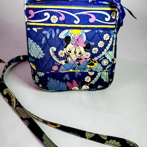 May include: A quilted crossbody bag in a deep blue fabric featuring a repeating floral pattern and a graphic print of Mickey and Minnie Mouse. The bag includes a zippered top closure, a front pocket, and a long adjustable shoulder strap.