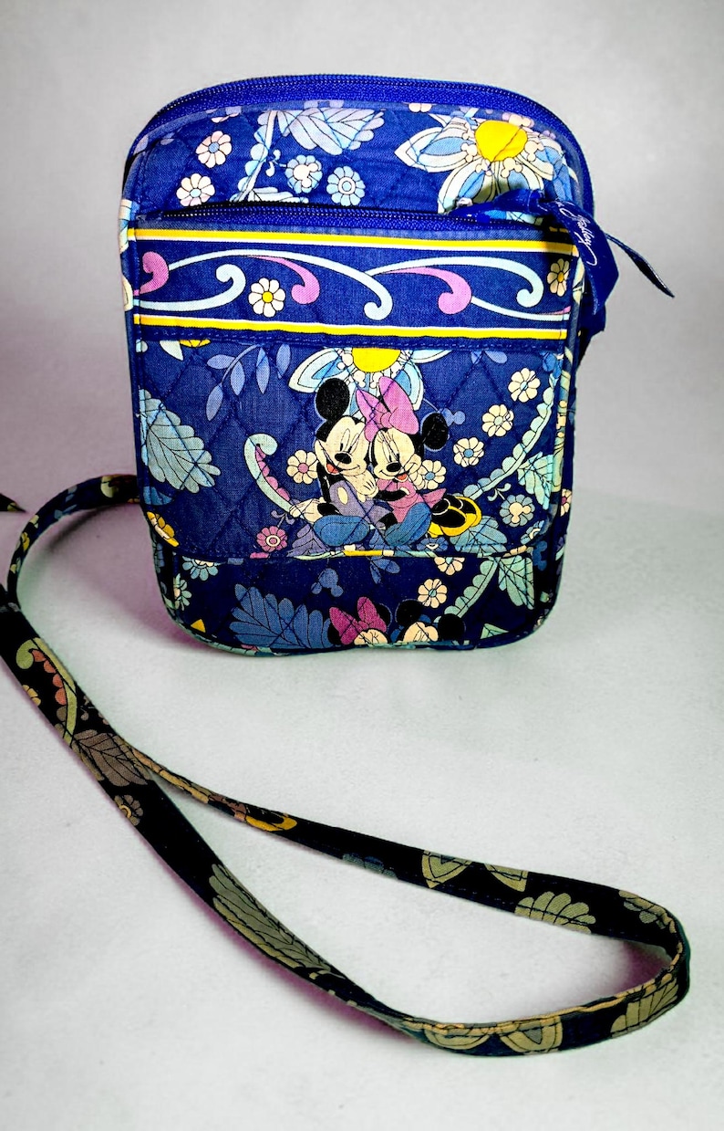 May include: A quilted crossbody bag in a deep blue fabric featuring a repeating floral pattern and a graphic print of Mickey and Minnie Mouse. The bag includes a zippered top closure, a front pocket, and a long adjustable shoulder strap.