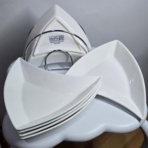 May include: A set of white, triangular-shaped plates with a matching metal rack. The plates are stacked and arranged on a white table. The plates have a modern, minimalist design, perfect for serving appetizers or desserts. The brand name "Living Colors" is visible.