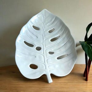 May include: A white ceramic leaf-shaped serving dish with a glossy finish. The dish features a detailed vein pattern and several oval cutouts, resembling a Monstera leaf. It sits on a wooden surface, suggesting a decorative or serving purpose.