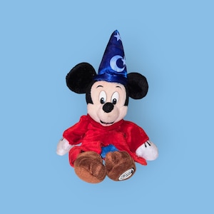 May include: A plush Mickey Mouse toy wearing a blue wizard hat with a crescent moon and star design. The toy is wearing a red robe and has a friendly expression.