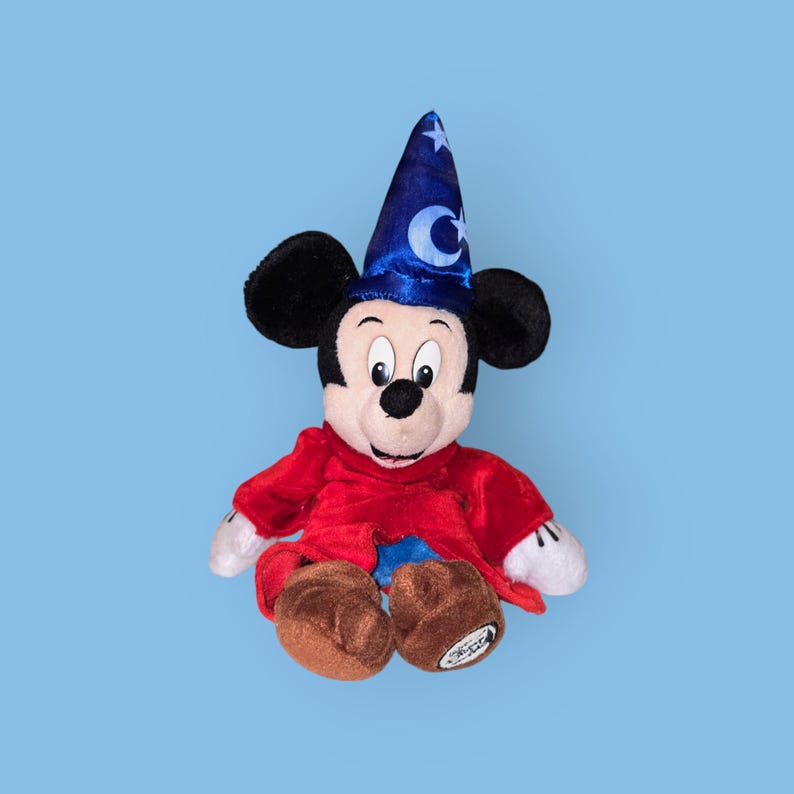 May include: A plush Mickey Mouse toy wearing a blue wizard hat with a crescent moon and star design. The toy is wearing a red robe and has a friendly expression.