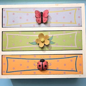 May include: A small wooden chest with three horizontal drawers in a white frame. The top drawer is light blue with a pink butterfly knob. The middle drawer is light green with a yellow flower knob. The bottom drawer is orange with a ladybug knob.