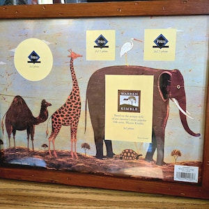 May include: A framed collage print featuring a camel, giraffe, and elephant. The print has a light blue background with yellow and brown accents. The frame is made of dark brown wood. The print includes the text "WARREN KIMBLE".