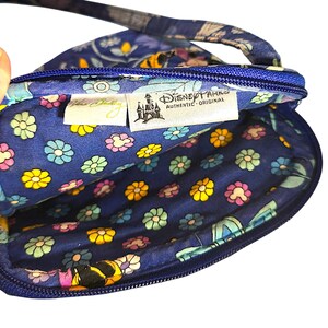 May include: A blue quilted crossbody bag with a floral pattern featuring small yellow, pink, and light blue flowers. The interior lining displays a matching floral print. Two fabric tags are visible inside, reading Vera Bradley and Disney Parks.