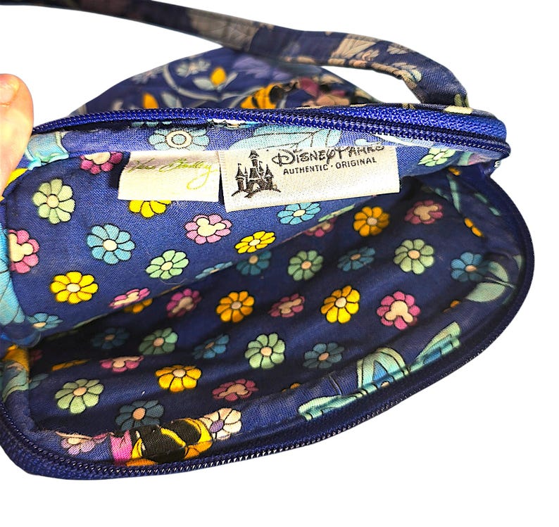 May include: A blue quilted crossbody bag with a floral pattern featuring small yellow, pink, and light blue flowers. The interior lining displays a matching floral print. Two fabric tags are visible inside, reading Vera Bradley and Disney Parks.