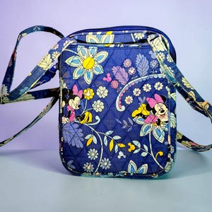 May include: A quilted crossbody bag in a deep blue fabric featuring a repeating pattern of Minnie Mouse, yellow flowers, and light blue foliage. The bag has a rectangular shape, a top zipper closure, and an adjustable matching fabric shoulder strap.