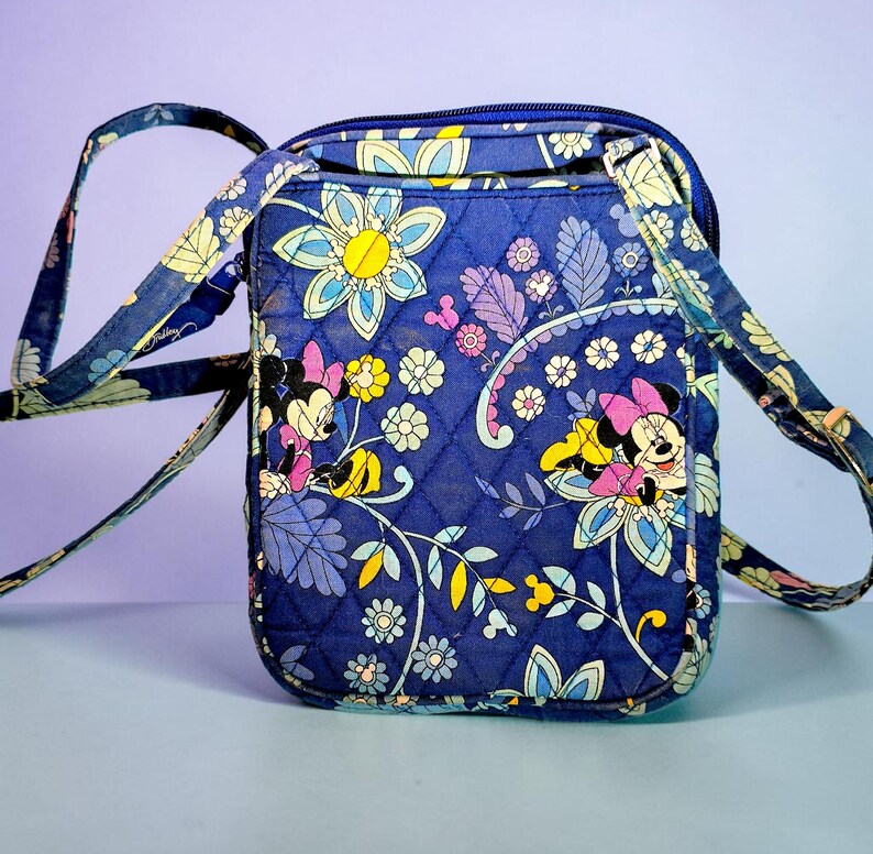 May include: A quilted crossbody bag in a deep blue fabric featuring a repeating pattern of Minnie Mouse, yellow flowers, and light blue foliage. The bag has a rectangular shape, a top zipper closure, and an adjustable matching fabric shoulder strap.